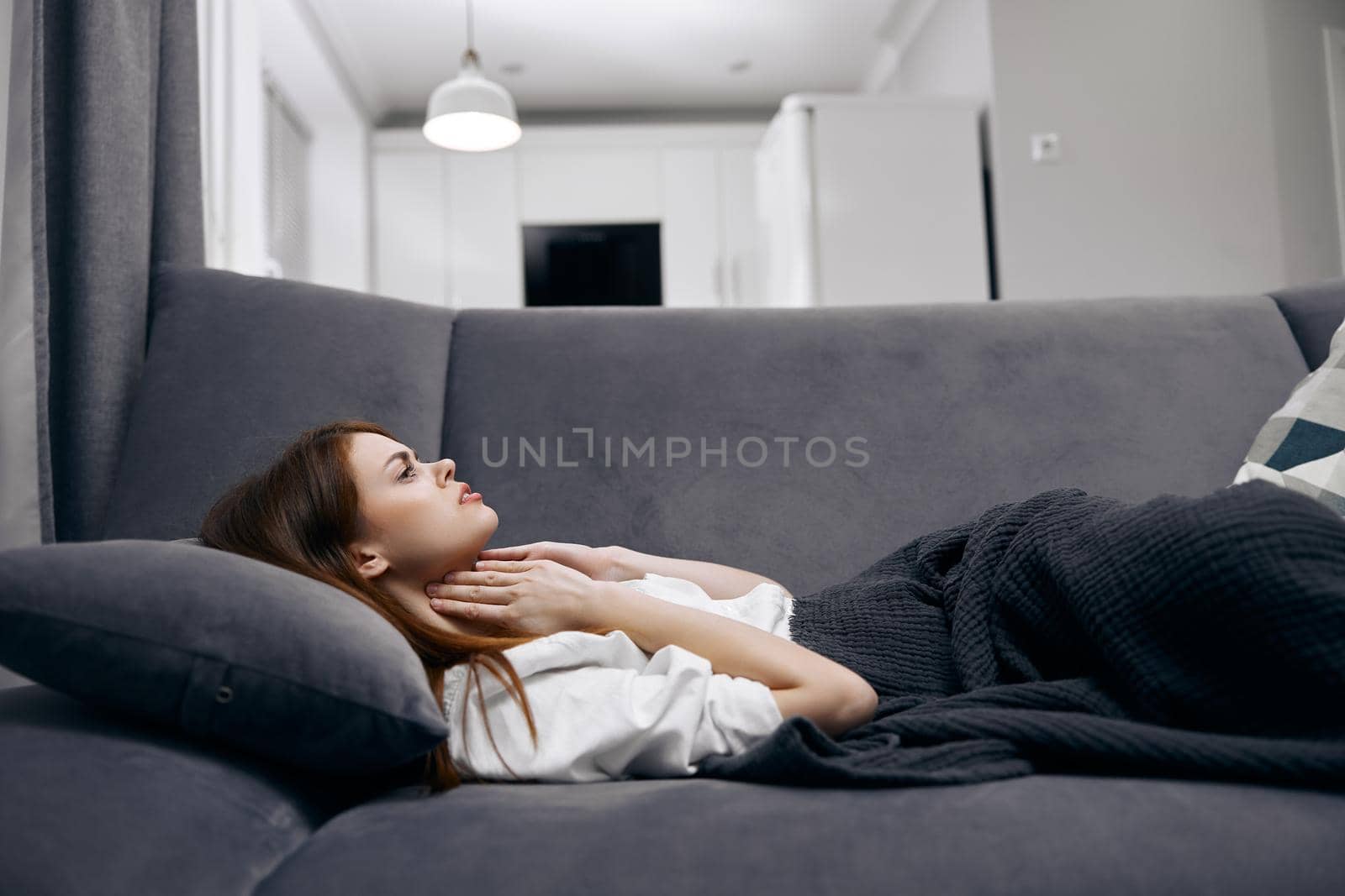 woman holding her neck while lying on the couch hiding with a blanket at home feeling unwell by SHOTPRIME