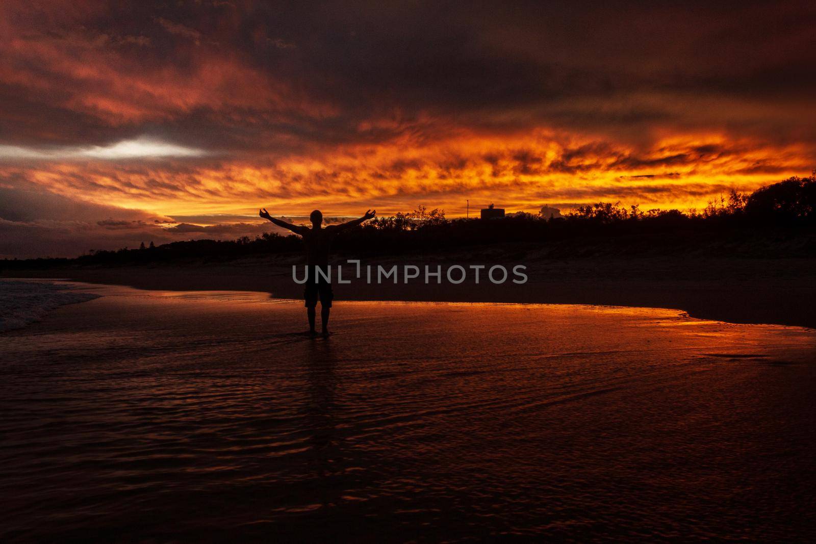 young man rising hands in the air because he is happy, with a colorfull sunset in the background at Noosaville beach, Sunshine Coast, Australia. by bettercallcurry