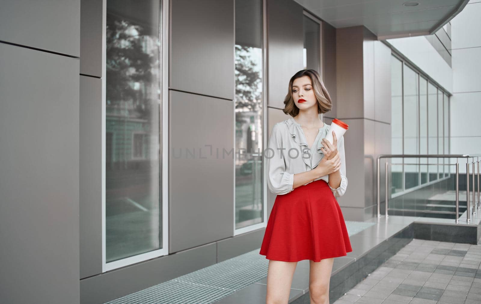 pretty blonde with a cup of coffee near the building on the street and red skirt white shirt by SHOTPRIME