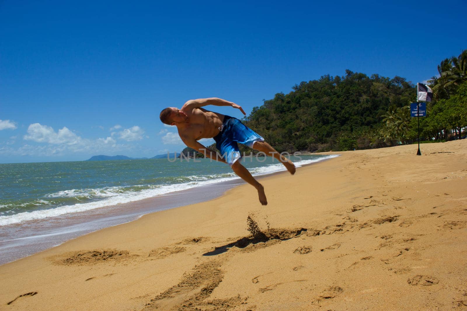 Muscular caucasian man doing a flip on the beach as ocean waves crash behind him, cape tribulation, queensland, australia by bettercallcurry