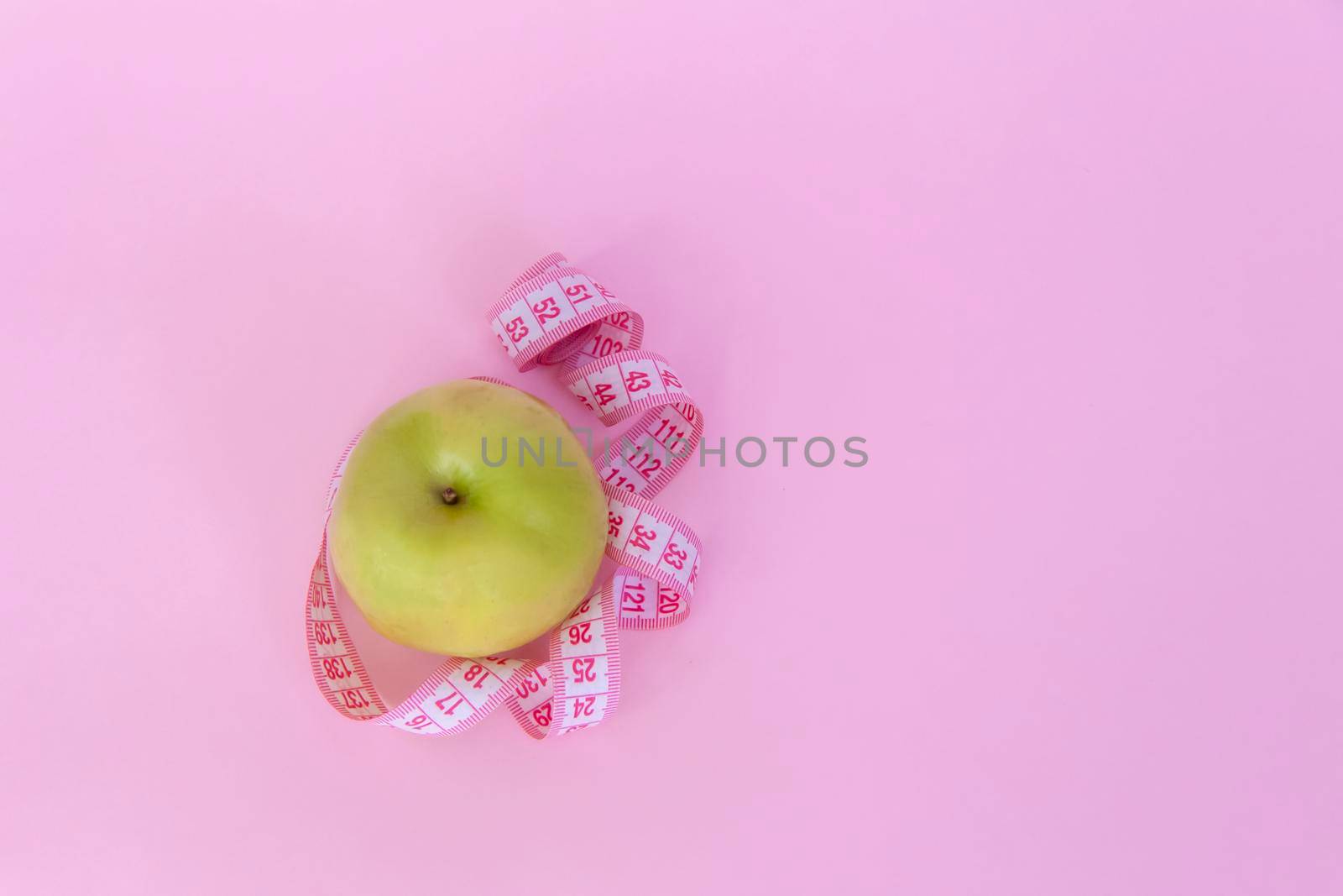 A green apple and a centimeter measuring tape on a pink background. Sports, sports nutrition, healthy eating, diet. Space for the text. Exercise, lose weight.