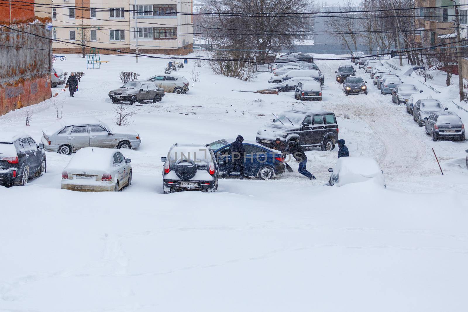 Tula, Russia - February 13, 2021: Two men pushing stuck Hyundai Solaris car through a snowy yard between rows of parked cars in deep snow in slope.