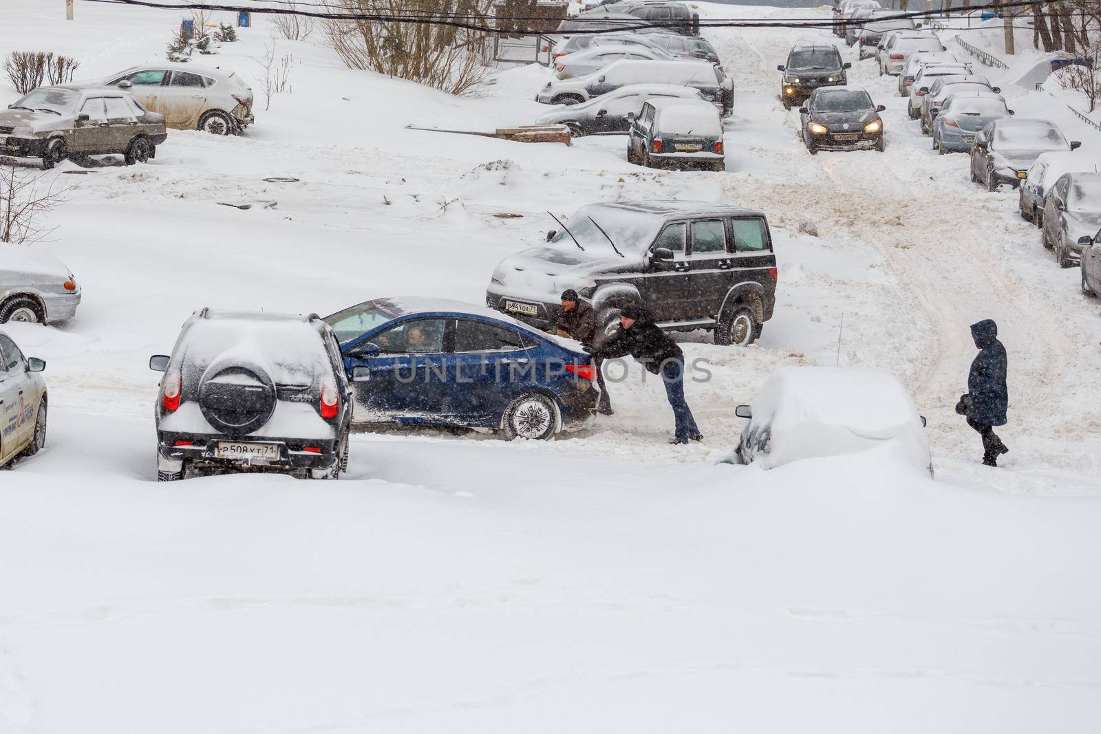 Two men pushing stuck Hyundai Solaris car through a snowy yard between rows of parked cars in deep snow in slope. by z1b