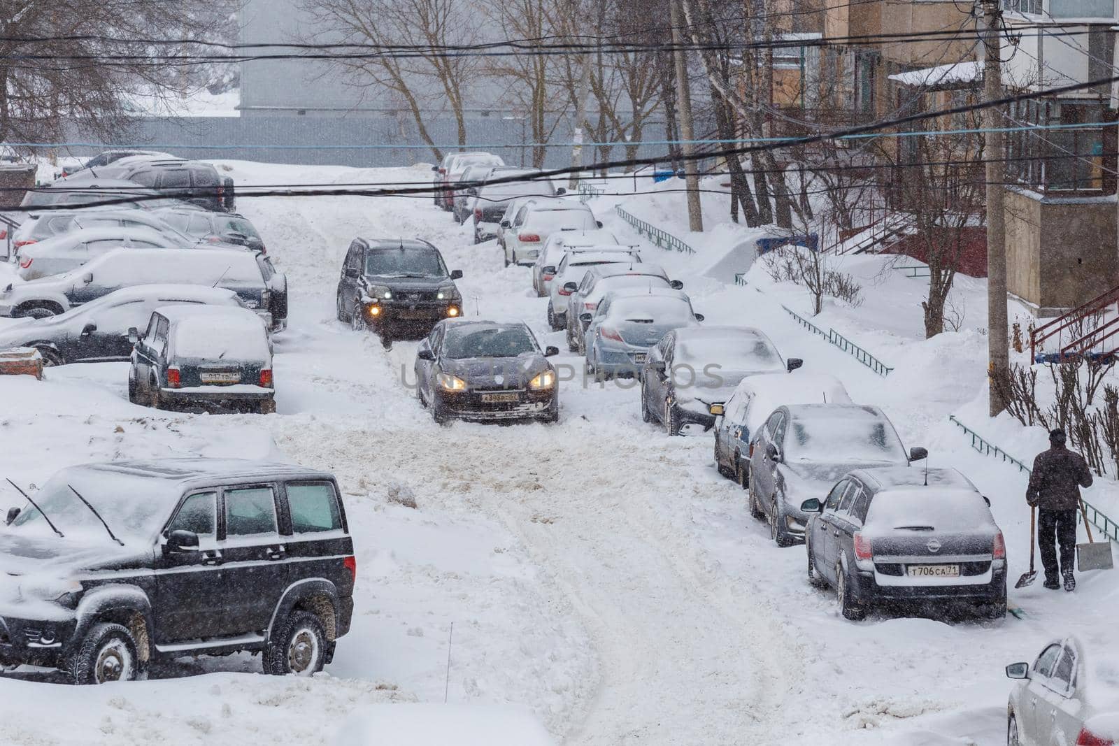 Two cars drive through a snowy yard between rows of parked cars in deep snow in Tula, Russia. by z1b