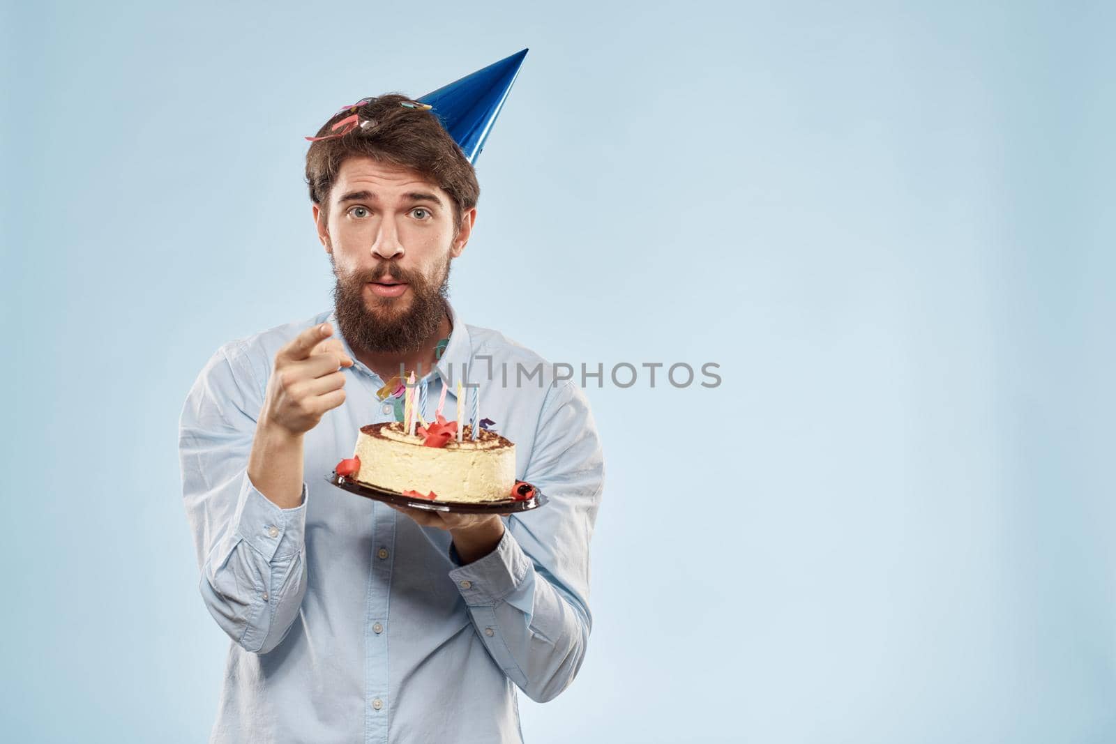 Bearded man with a plate of cake on a blue background and a birthday party hat on his head. High quality photo