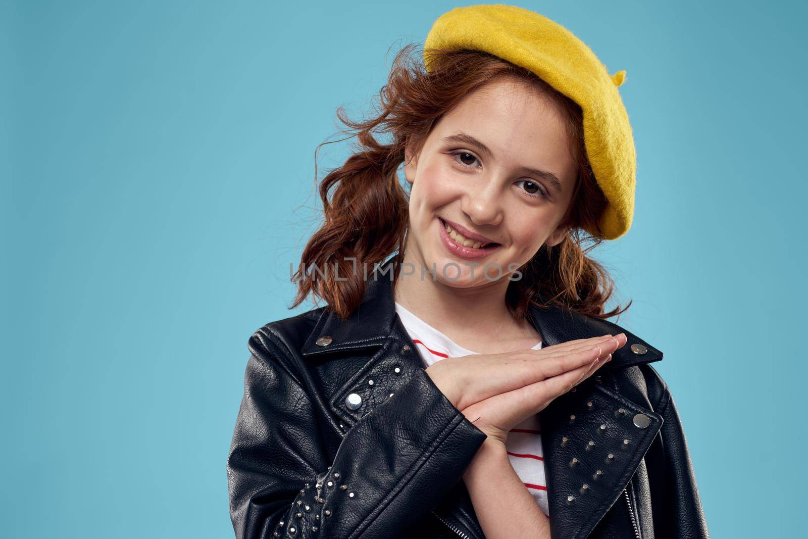 A fashionable girl in a leather jacket and a yellow hat on a blue background gestures with her hands emotions. High quality photo