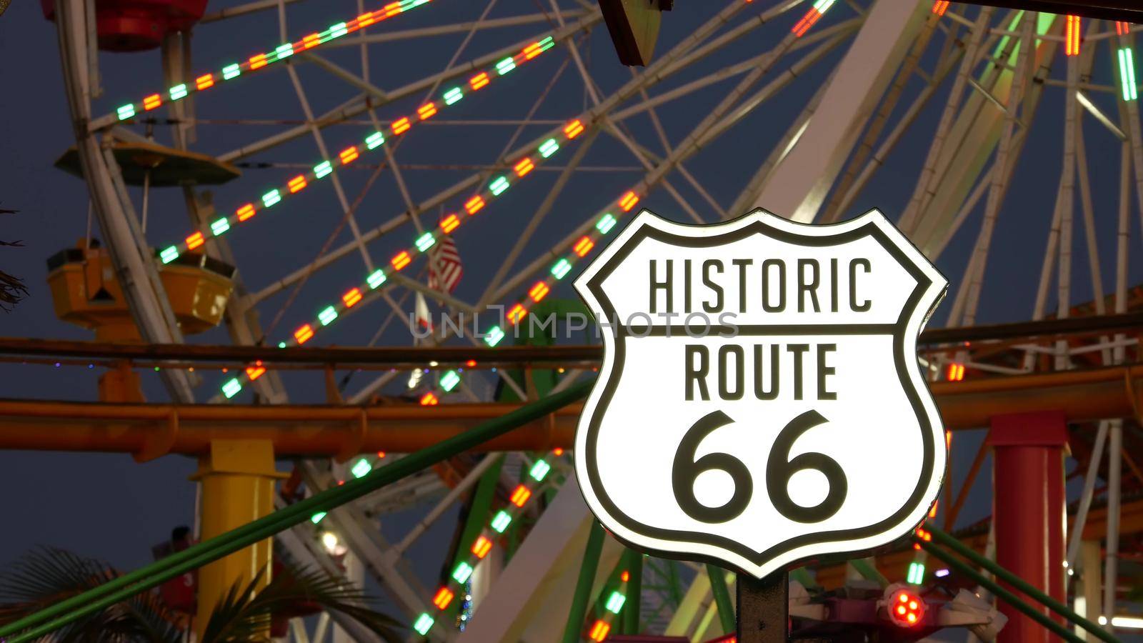 SANTA MONICA, LOS ANGELES, USA - 28 OCT 2019: Iconic road sign glowing, historic route 66. Famous california symbol, pier of pacific ocean resort. Illuminated festive ferris wheel in amusement park.