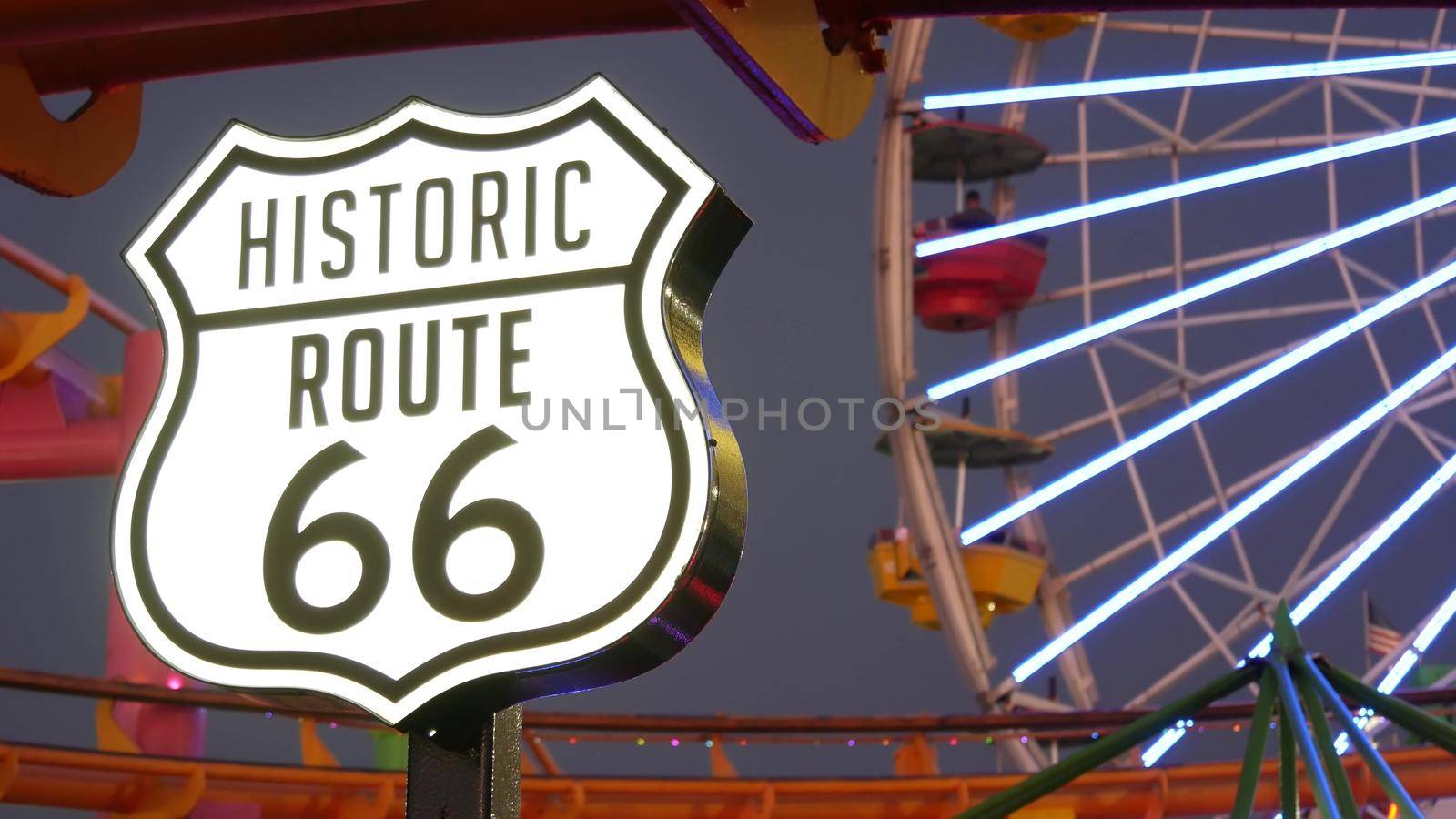 SANTA MONICA, LOS ANGELES, USA - 28 OCT 2019: Iconic road sign glowing, historic route 66. Famous california symbol, pier of pacific ocean resort. Illuminated festive ferris wheel in amusement park.