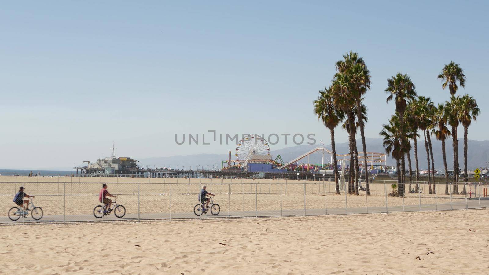 SANTA MONICA, LOS ANGELES CA USA - 28 OCT 2019: California summertime beach aesthetic, people walking and ride cycles on bicycle path. Amusement park on pier and palms. American pacific ocean resort.