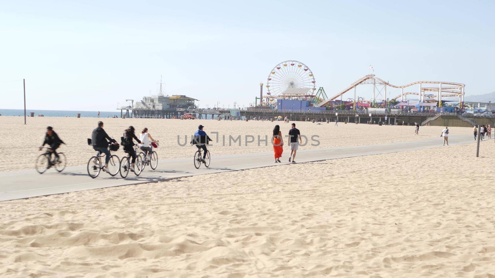 SANTA MONICA, LOS ANGELES CA USA - 28 OCT 2019: California summertime beach aesthetic, people walking and ride cycles on bicycle path. Amusement park on pier and palms. American pacific ocean resort by DogoraSun