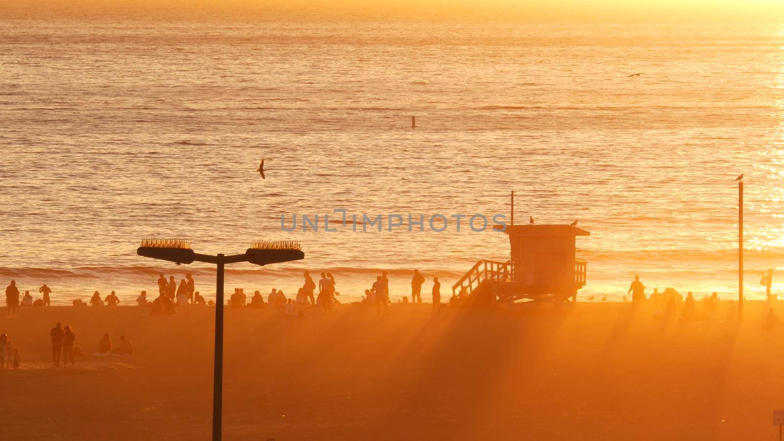 SANTA MONICA, LOS ANGELES, USA - 28 OCT 2019: California summertime beach aesthetic, atmospheric golden sunset. Unrecognizable people silhouettes, sun rays over pacific ocean waves. Lifeguard tower by DogoraSun