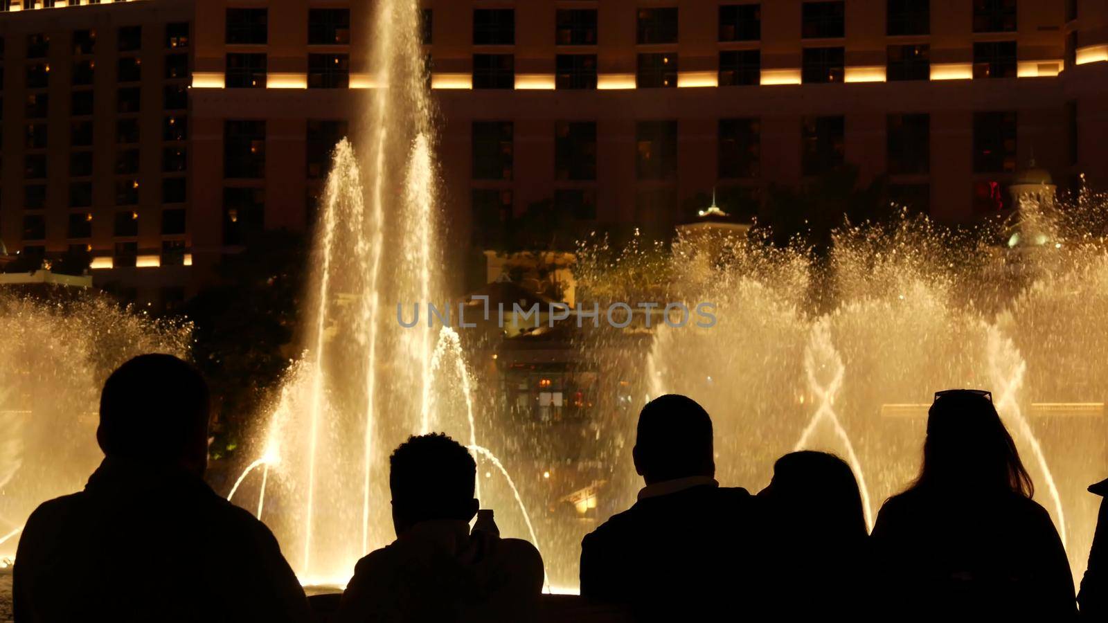 LAS VEGAS, NEVADA USA - 13 DEC 2019: People looking at Bellagio fountain musical performance at night. Contrast silhouettes and glowing dancing splashing water. Entertainment show in gambling city by DogoraSun