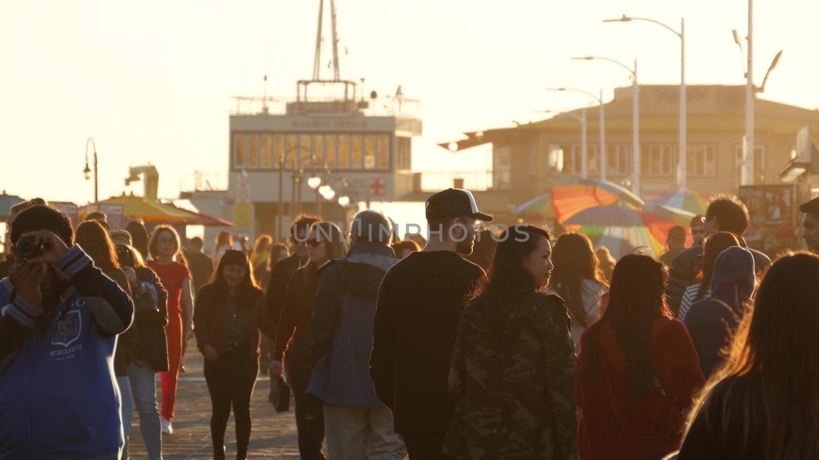 SANTA MONICA, LOS ANGELES CA USA - 19 DEC 2019: Many multiracial people walking on pier. Pedestrians walk on overcrowded seafront promenade. Crowd in golden sun light on broadwalk, sun rays over heads by DogoraSun