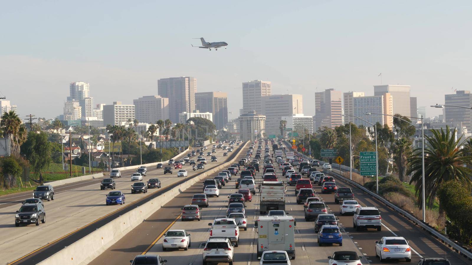 SAN DIEGO, CALIFORNIA USA - 15 JAN 2020: Busy intercity freeway, traffic jam on highway during rush hour. Urban skyline, highrise skyscraper and landing plane. Flying airplane, transportation concept by DogoraSun