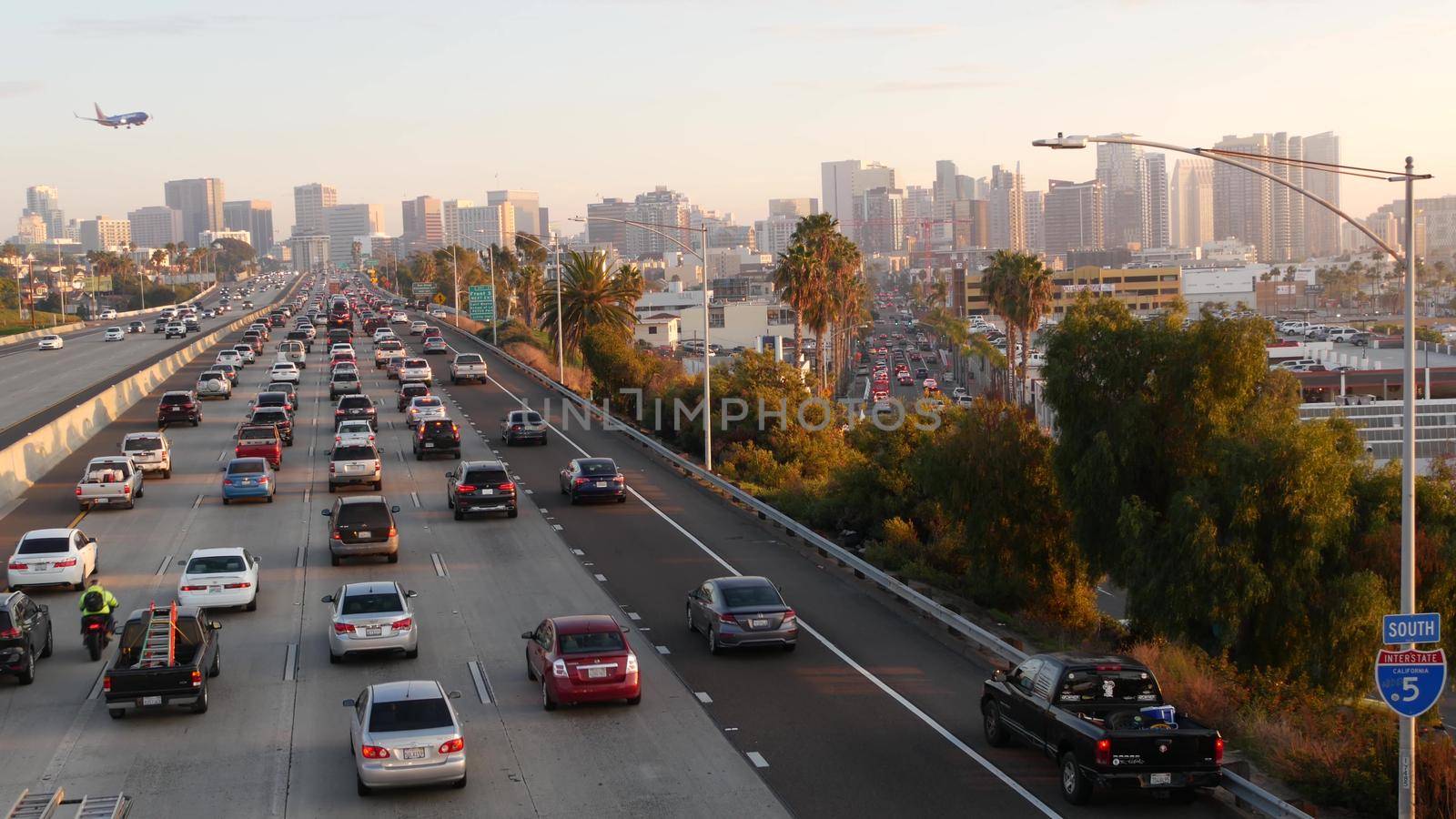 SAN DIEGO, CALIFORNIA USA - 15 JAN 2020: Busy intercity freeway, traffic jam on highway during rush hour. Urban skyline, highrise skyscraper and landing plane. Flying airplane, transportation concept by DogoraSun