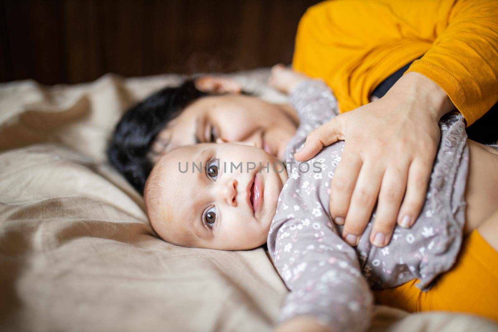 Cute baby lying down on bed next to her sleeping mother. Young woman wearing yellow clothing on bed and tenderly hugging adorable baby. Lovely maternity