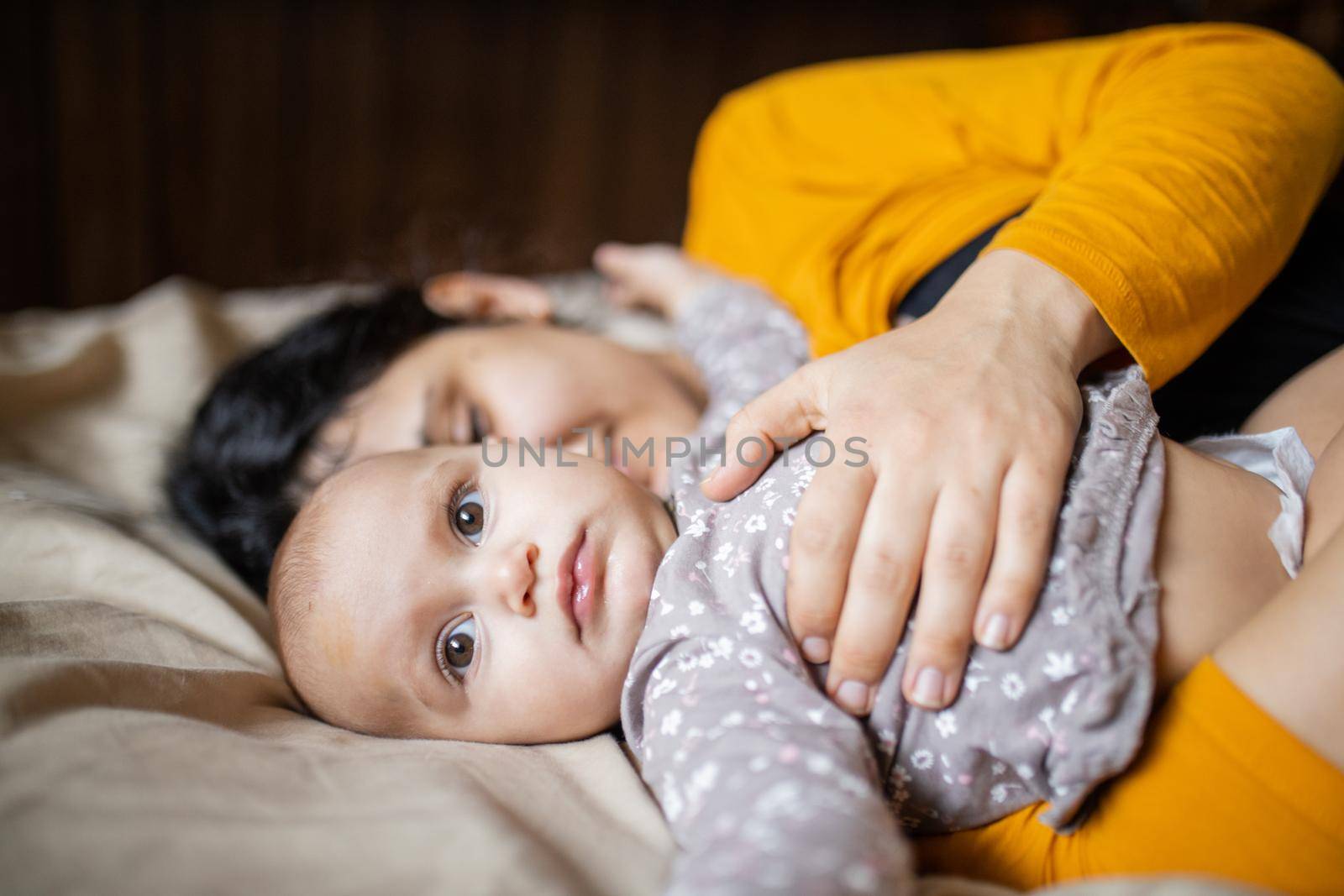 Cute baby lying down on bed next to her sleeping mother. Young woman wearing yellow clothing on bed and tenderly hugging adorable baby. Lovely maternity