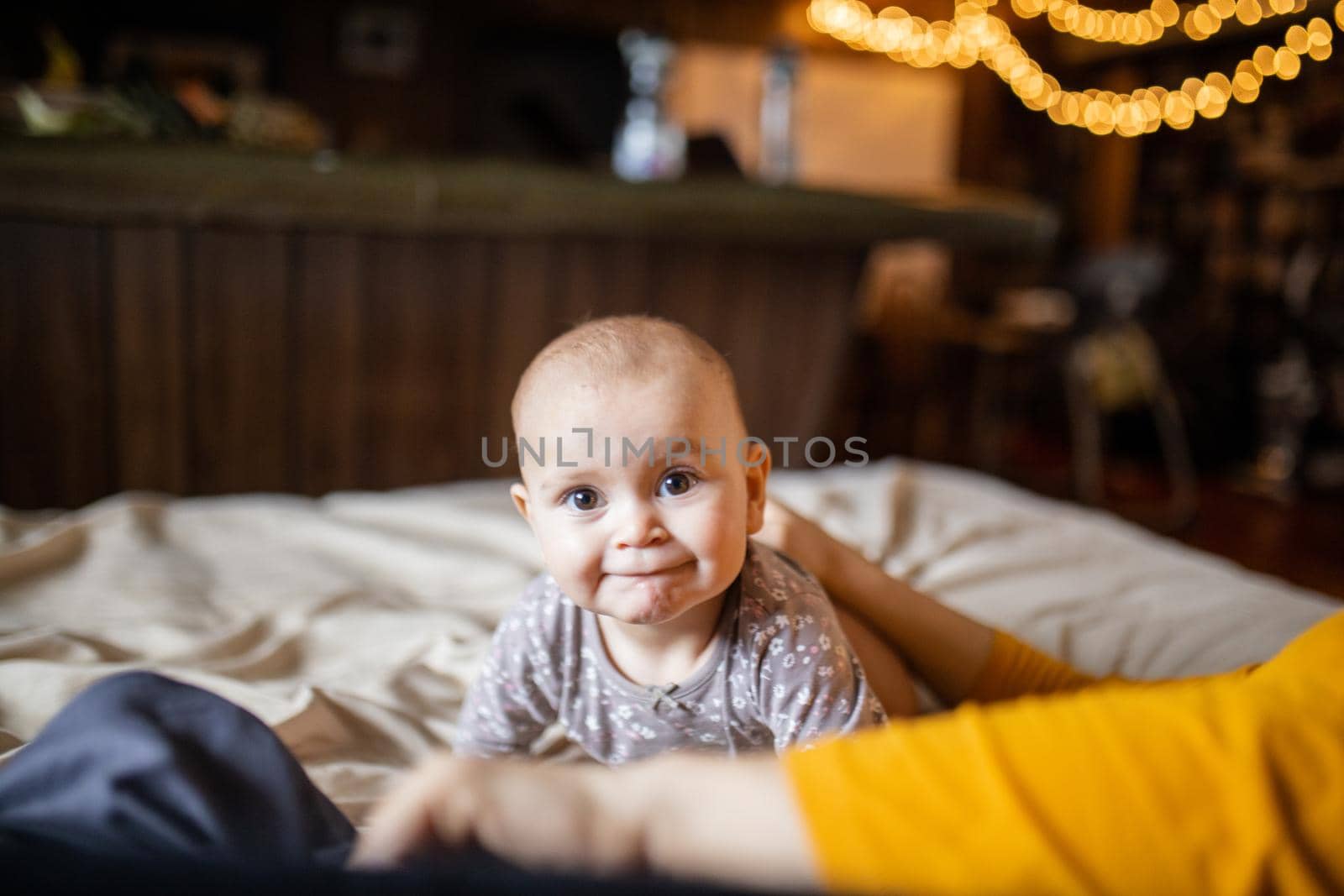 Adorable and curious-looking baby lying on her stomach above bed with blurry background. Lovely baby on bed with cream color sheets next to her mother. Cute toddlers