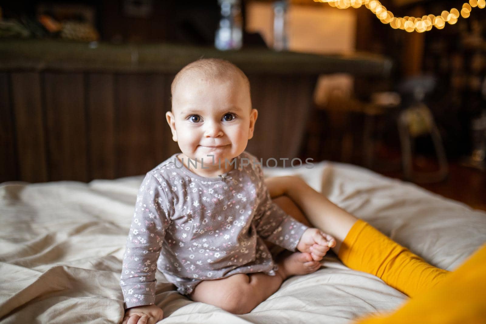 Adorable and happy baby sitting on bed with blurry background. Lovely baby on bed with cream color sheets next to her mother. Cute toddlers