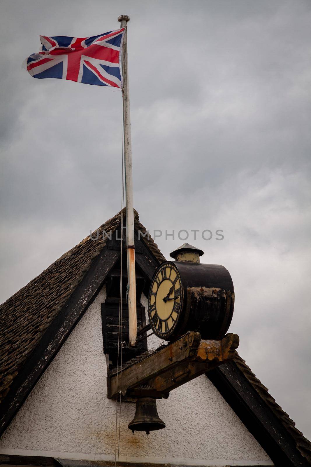 Shot of a flagpole in the top of a roof with the Union Jack by an old clock against a cloudy grey sky