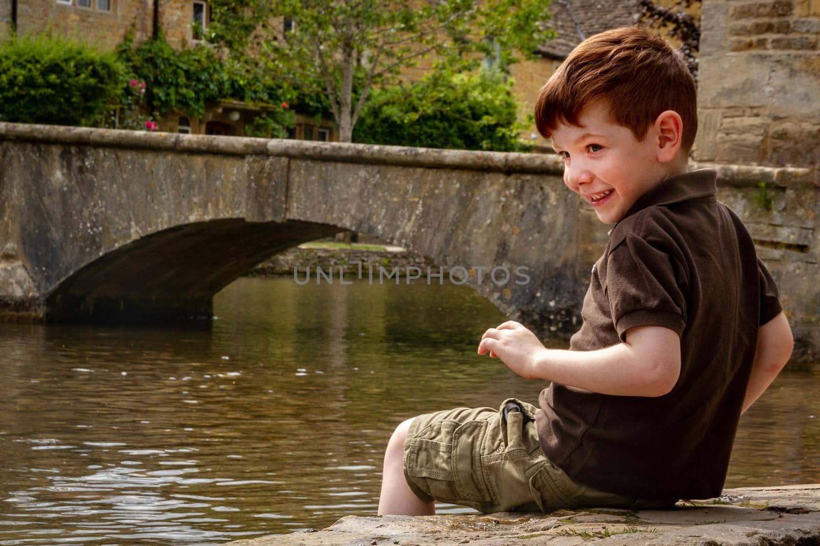 Portrait of a cute, red-haired, blue-eyed, four years old boy on brown shirt and shorts, sitting by a river with his feet on the water and a stone bridge in the background