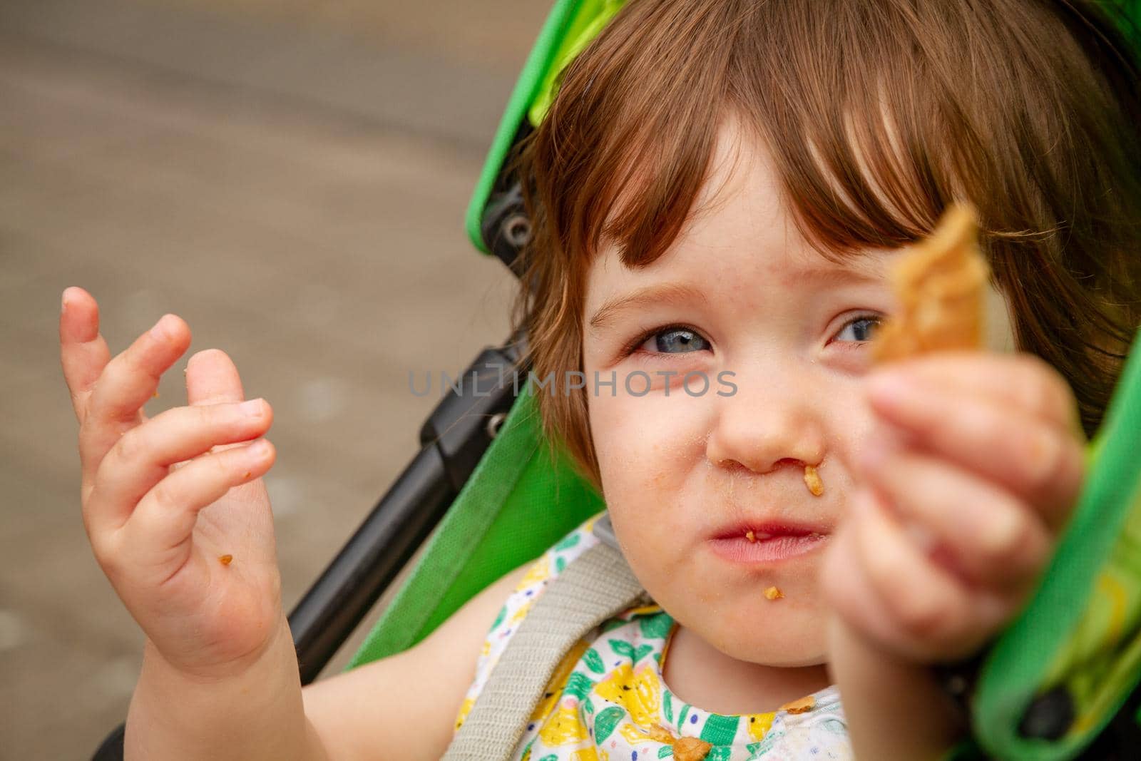 Portrait of a cute, blue-eyed, brown-haired baby girl sitting in a green pushchair holding a piece of waffle towards the camera while eating