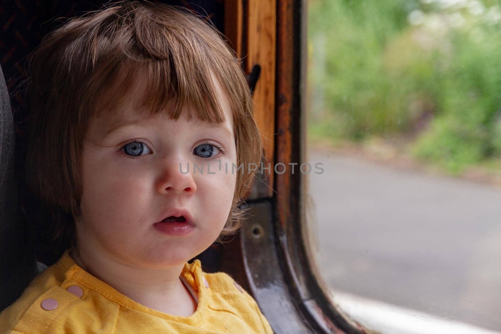 Portrait of a cute, blue-eyed, brown-haired baby girl in a yellow shirt sitting on a window