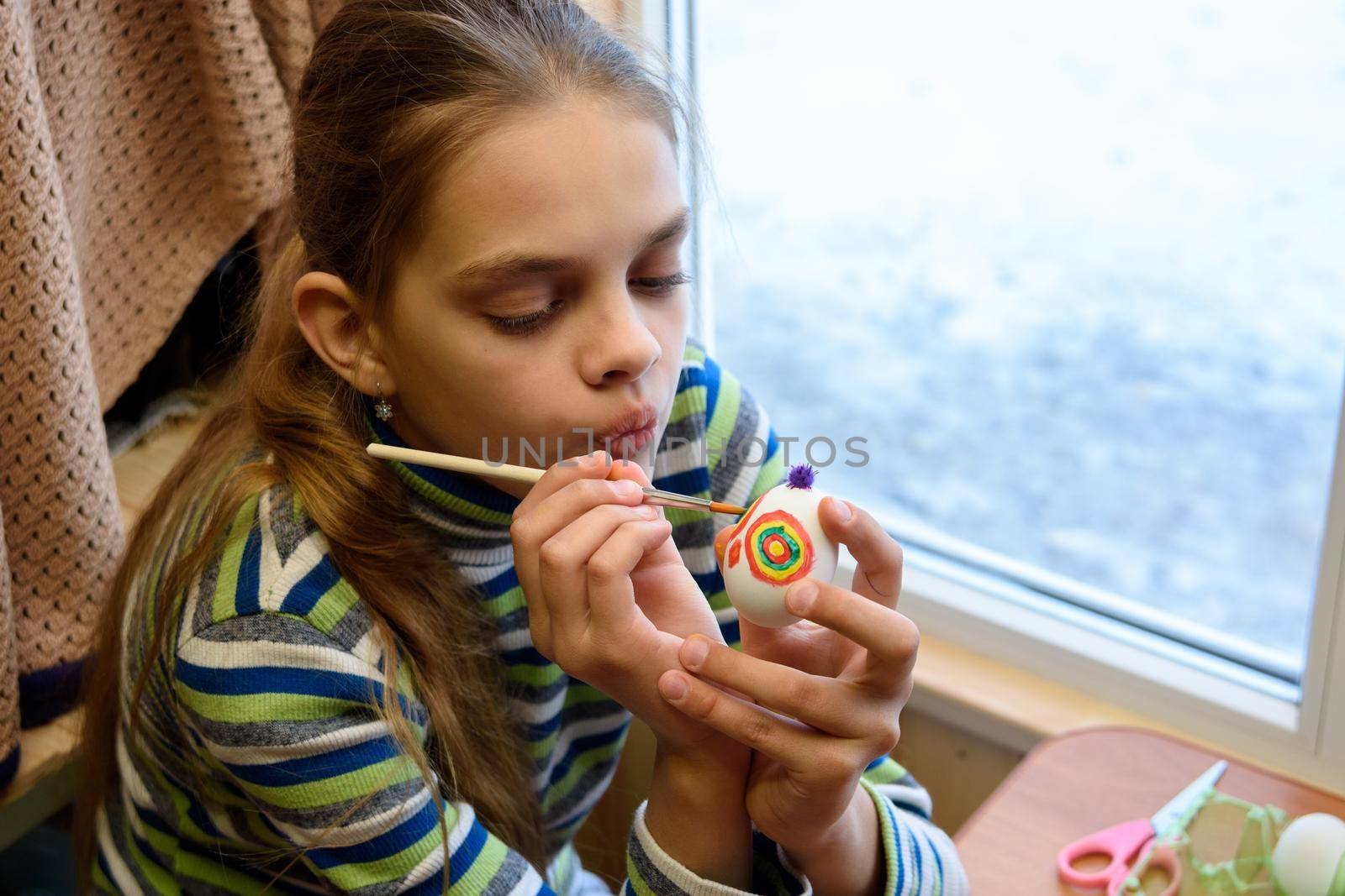 Girl paints Easter eggs while sitting at the table by the window