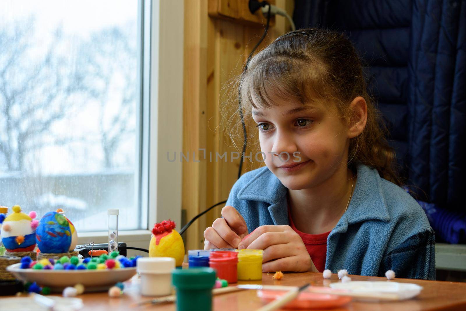 The girl funny thought painting Easter eggs while sitting at the table by the window