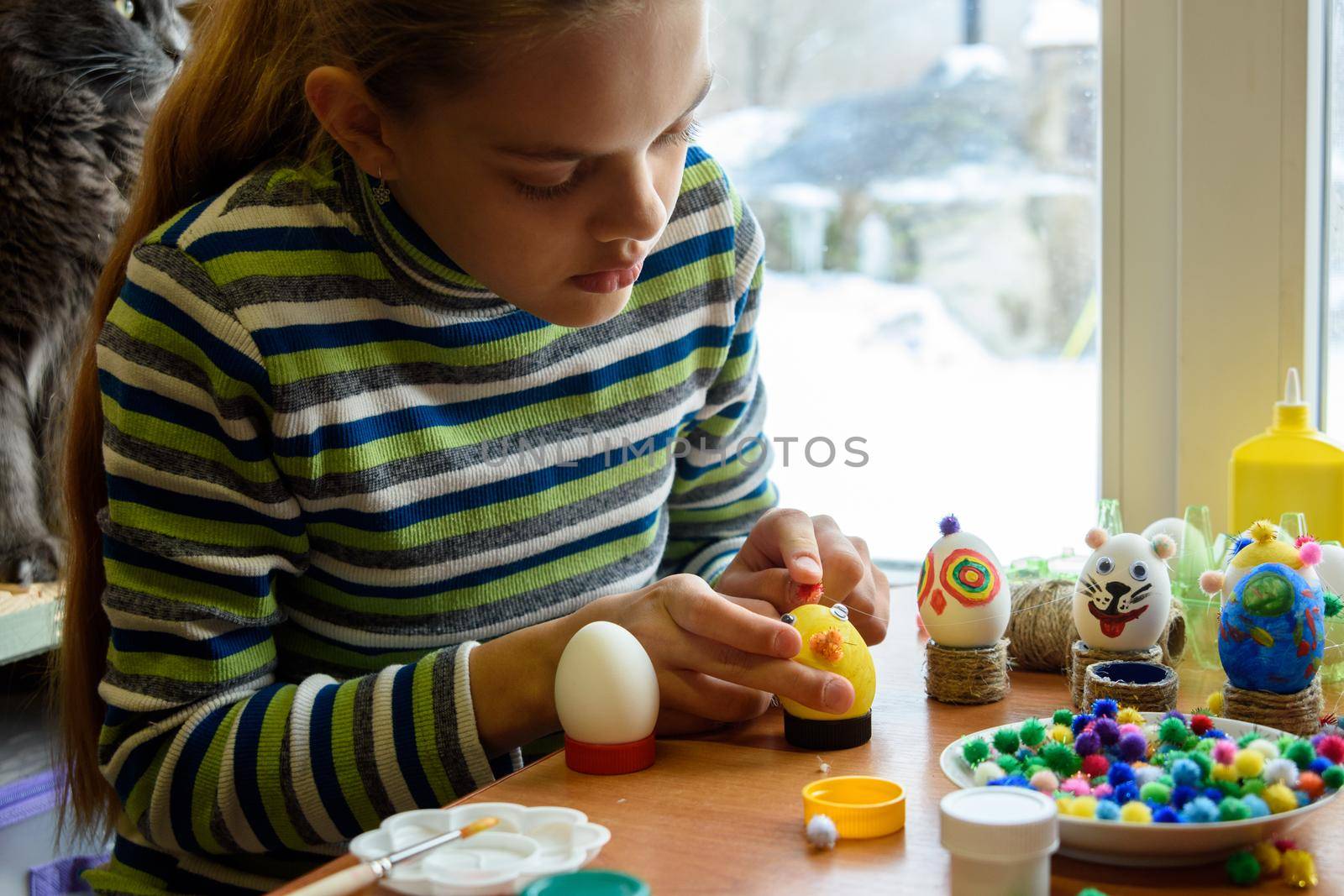 A girl sits at a table by the window and paints Easter eggs with a brush and paints