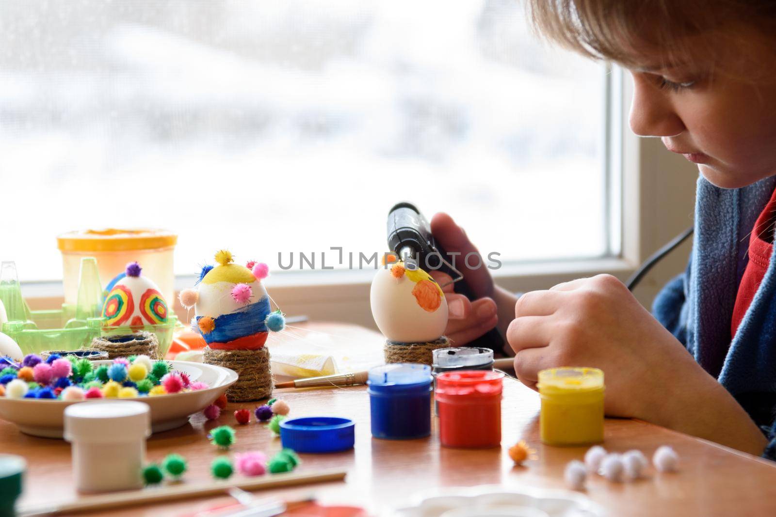 A girl glues elements to an Easter egg with a glue gun