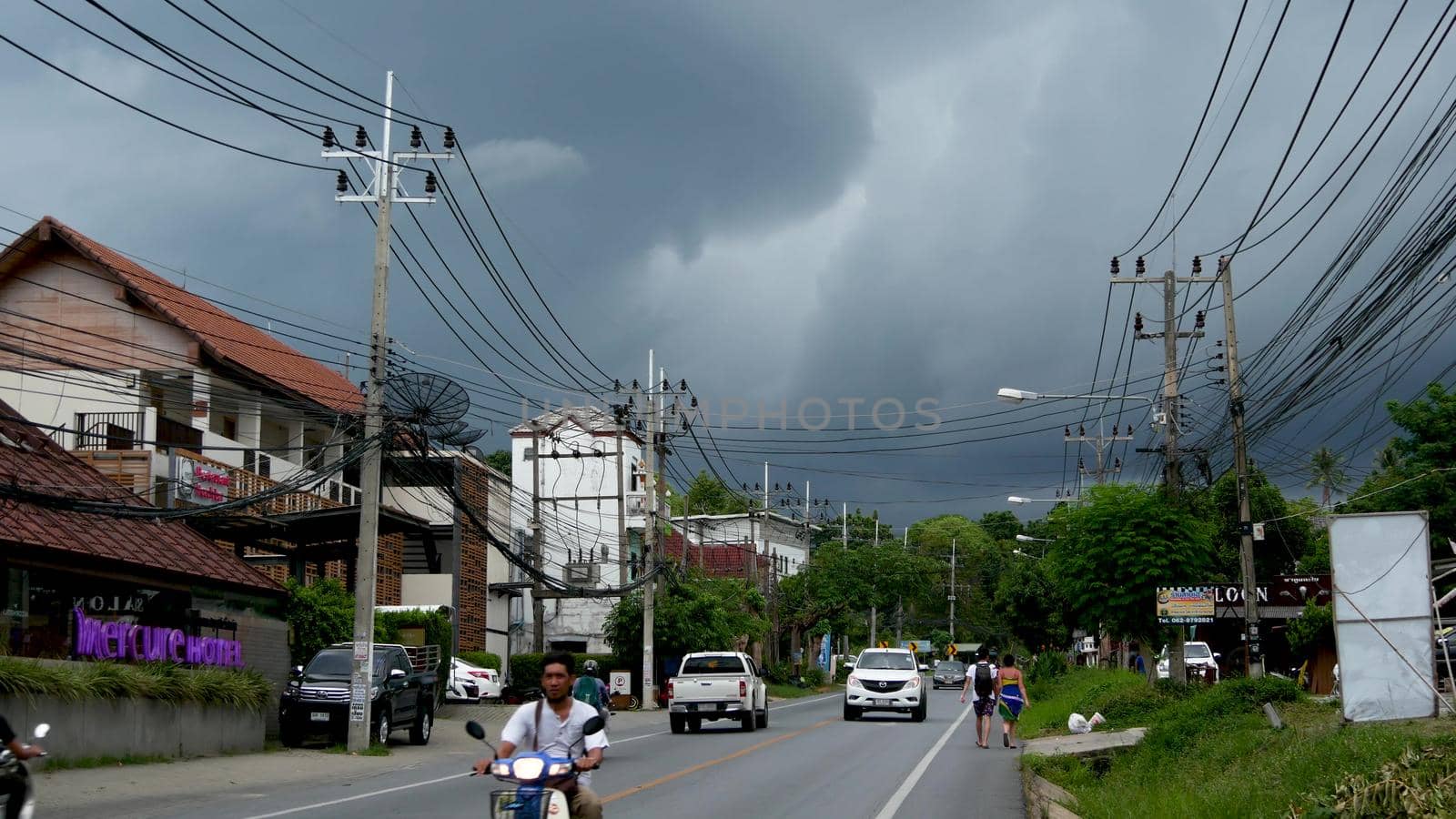 KOH SAMUI ISLAND, THAILAND - 11 JULY 2019 Busy transport populated city street in cloudy day. Typical street full of motorcycles and cars. Thick blue clouds before storm during wet season. by DogoraSun
