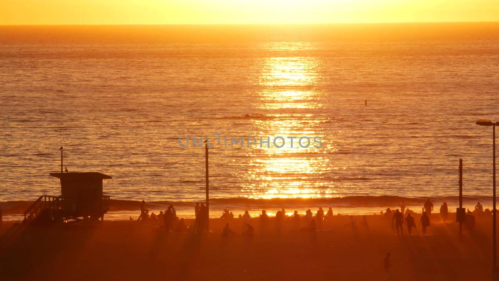 SANTA MONICA, LOS ANGELES, USA - 28 OCT 2019: California summertime beach aesthetic, atmospheric golden sunset. Unrecognizable people silhouettes, sun rays over pacific ocean waves. Lifeguard tower.