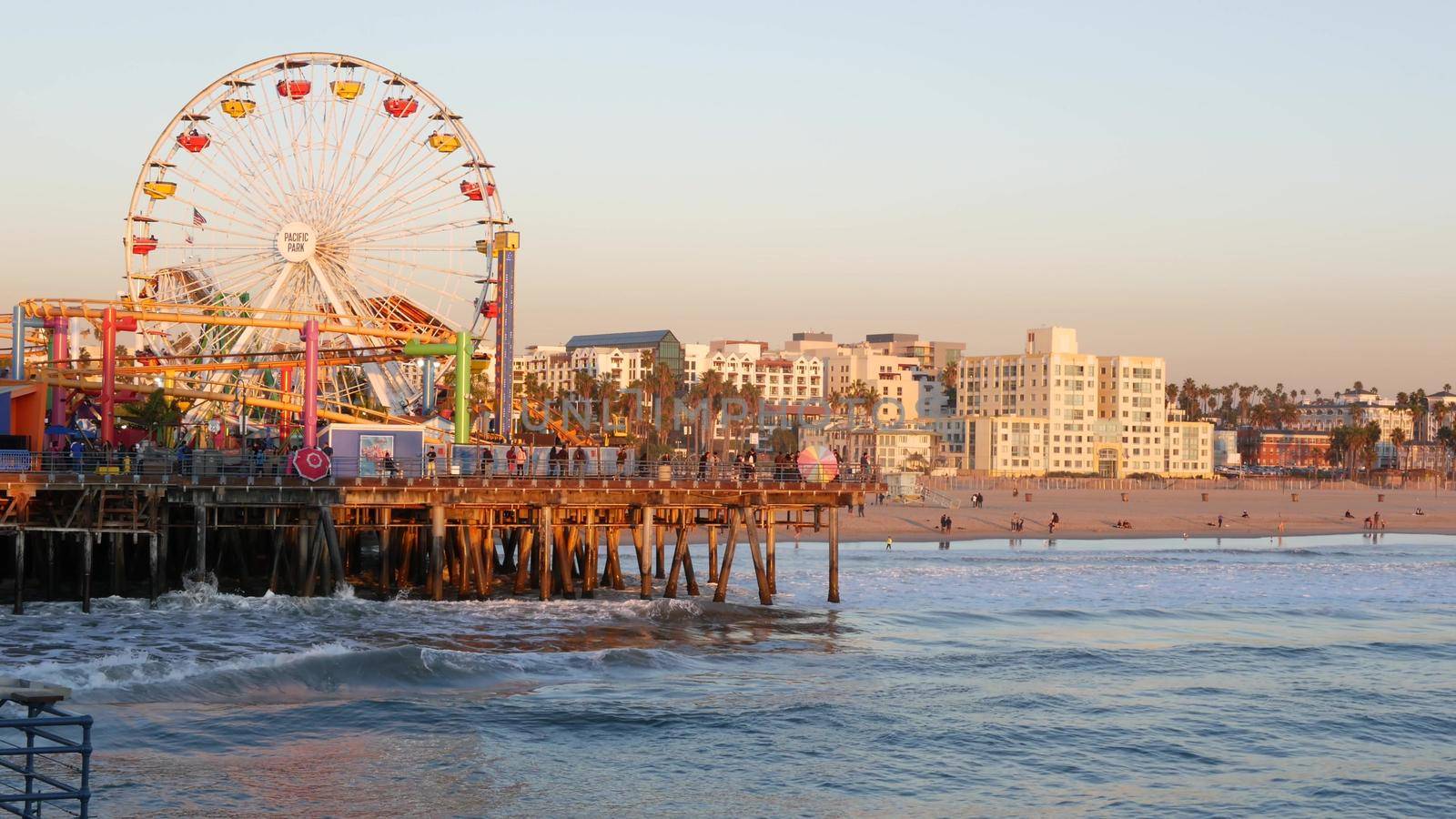 SANTA MONICA, LOS ANGELES CA USA - 19 DEC 2019: Classic ferris wheel in amusement park on pier. California summertime beach aesthetic, ocean waves in pink sunset. Summertime iconic symbol by DogoraSun