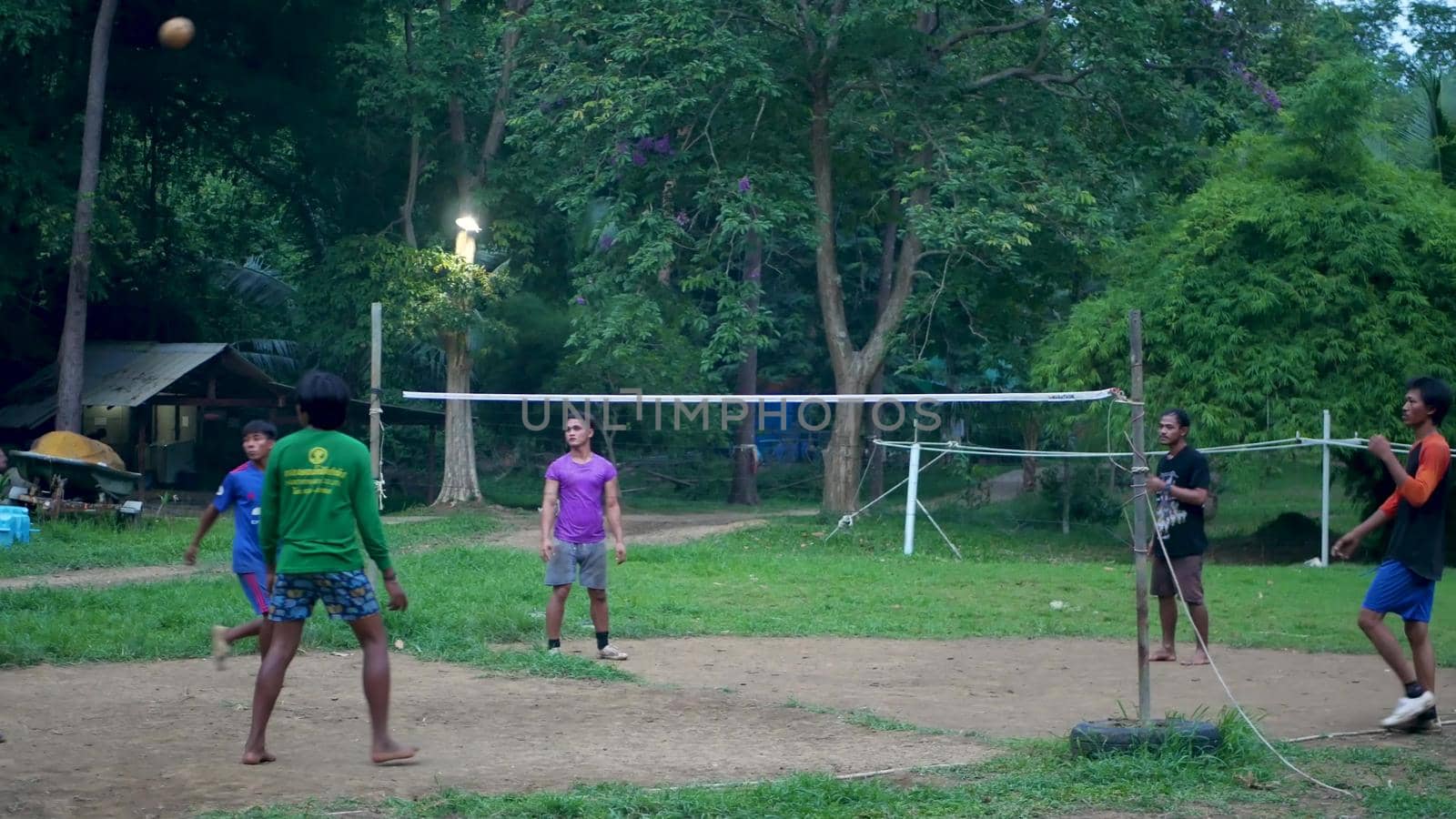 ANG THONG, THAILAND - 9 JUNE 2019: Thai teenagers playing sepak takraw in park. Group of men playing kick volleyball against green trees in yard in local settlement. Traditional national sport game by DogoraSun