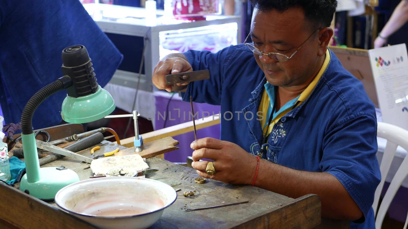 BANGKOK, THAILAND - 10 JULY, 2019: Ethnic jeweler working during exhibition. Ethnic craftsman in glasses sitting at shabby table and making jewelry while taking part in trade show.