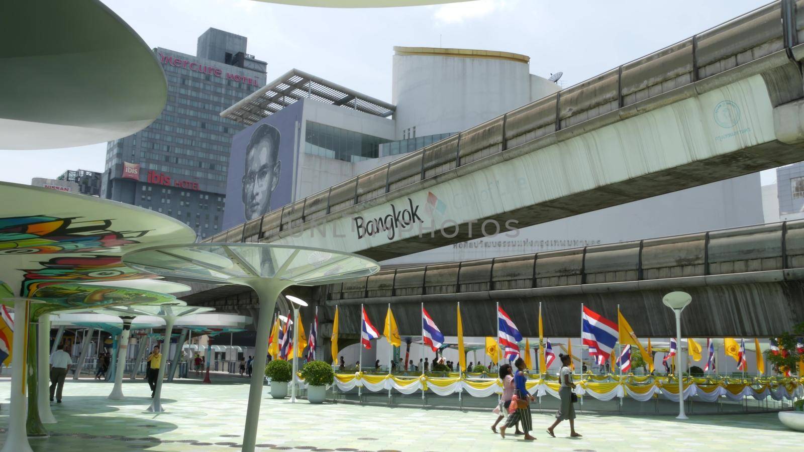 BANGKOK, THAILAND - 11 JULY, 2019: Pedestrians walking on the bridge near MBK and Siam Square under BTS train line. People in festive modern city decorated with waving national flags and royal symbol by DogoraSun