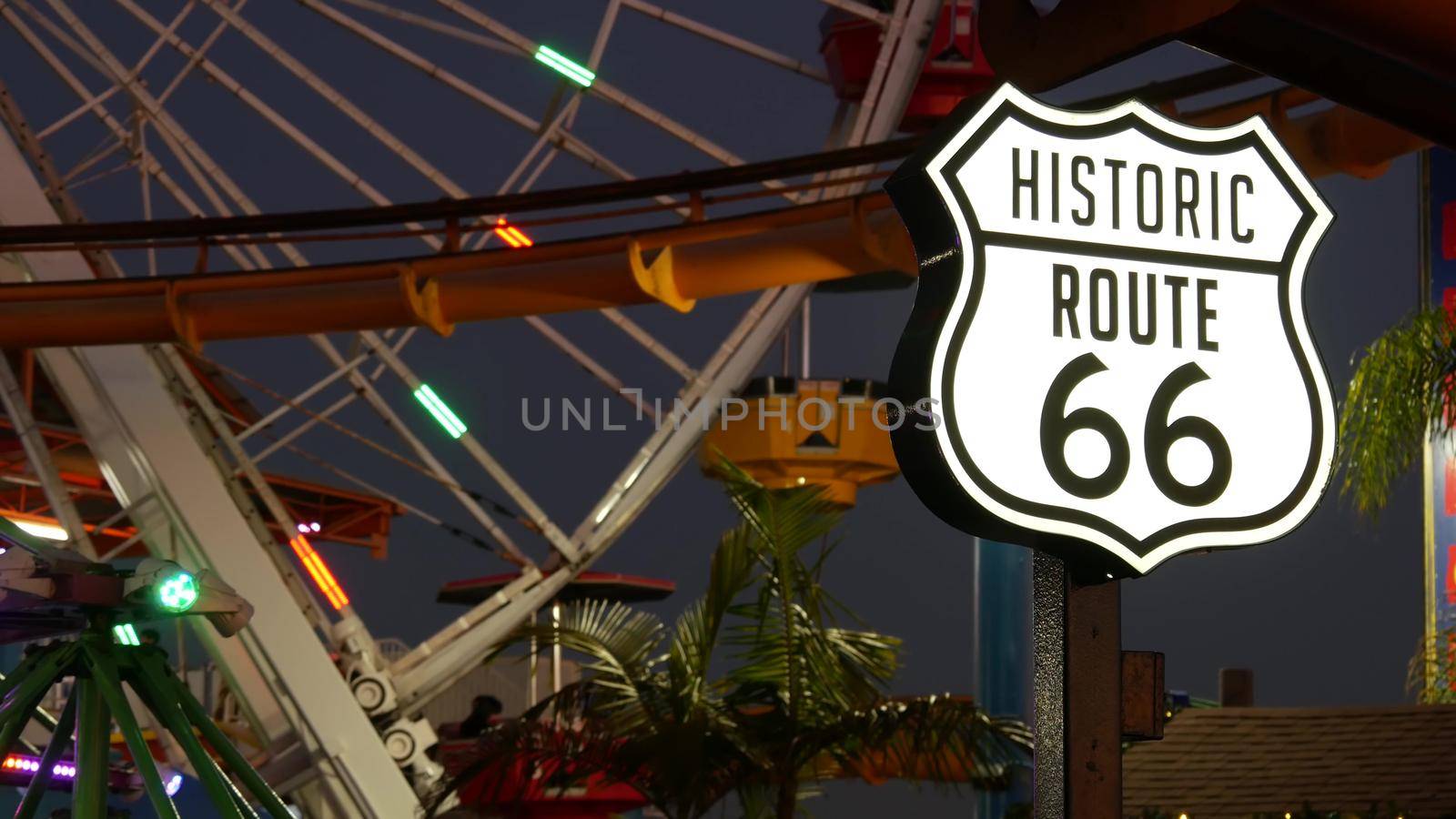 SANTA MONICA, LOS ANGELES, USA - 28 OCT 2019: Iconic road sign glowing, historic route 66. Famous california symbol, pier of pacific ocean resort. Illuminated festive ferris wheel in amusement park.