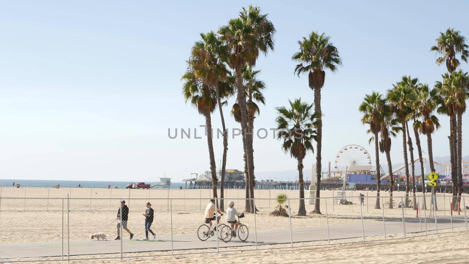 SANTA MONICA, LOS ANGELES CA USA - 28 OCT 2019: California summertime beach aesthetic, people walking and ride cycles on bicycle path. Amusement park on pier and palms. American pacific ocean resort.