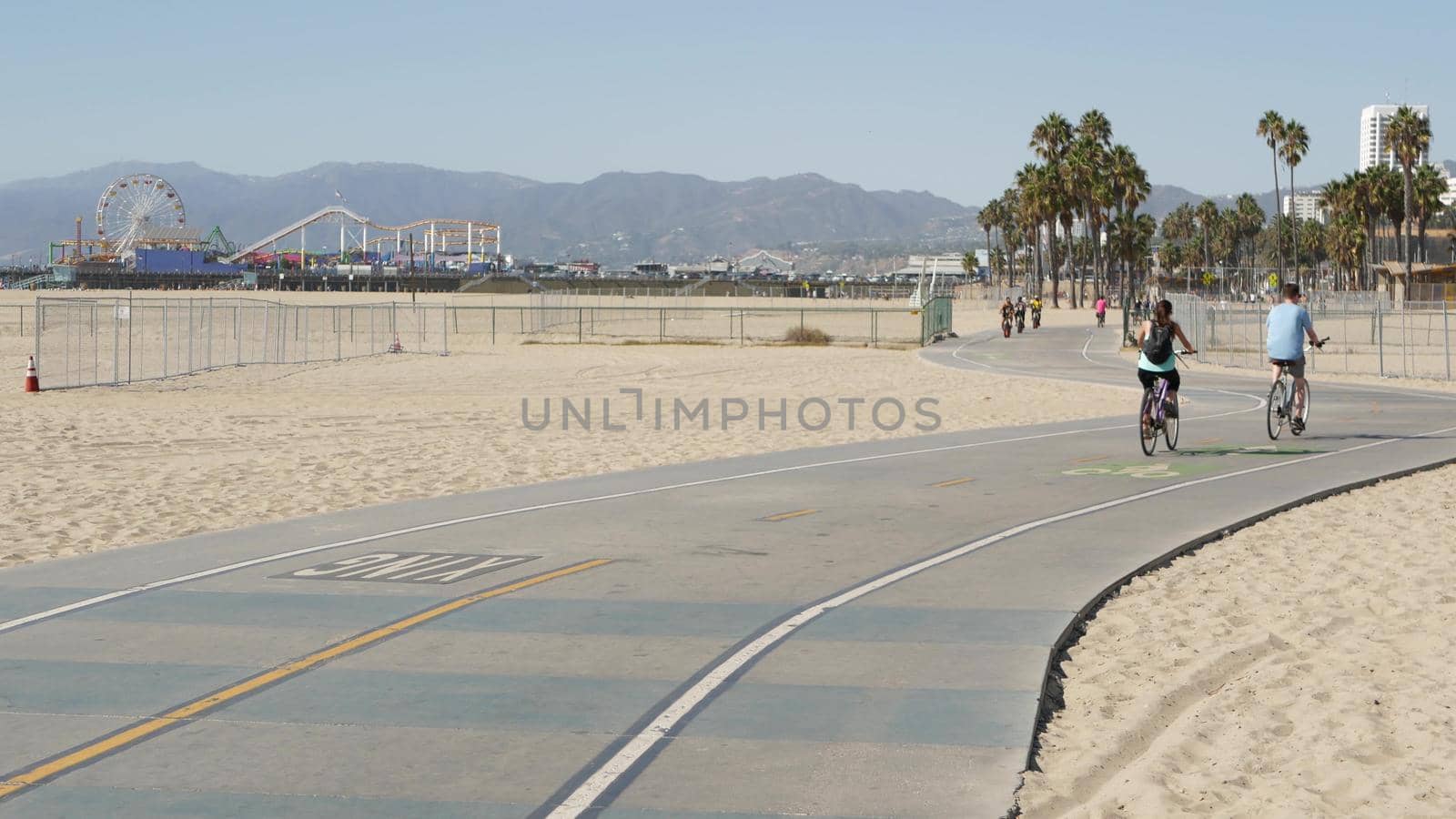 SANTA MONICA, LOS ANGELES CA USA - 28 OCT 2019: California summertime beach aesthetic, people walking and ride cycles on bicycle path. Amusement park on pier and palms. American pacific ocean resort by DogoraSun