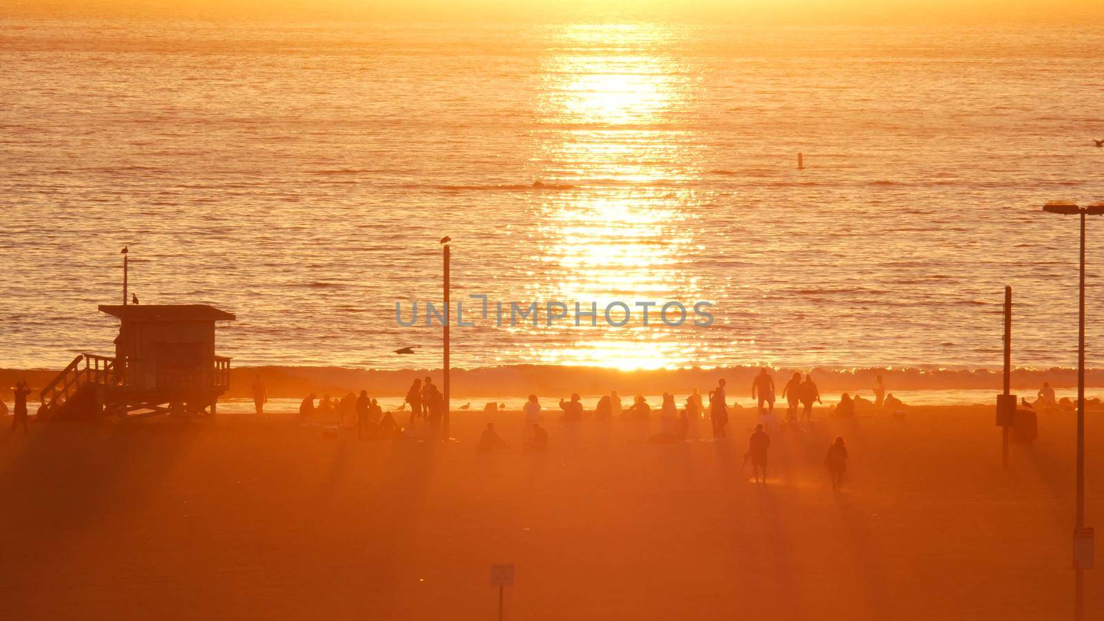 SANTA MONICA, LOS ANGELES, USA - 28 OCT 2019: California summertime beach aesthetic, atmospheric golden sunset. Unrecognizable people silhouettes, sun rays over pacific ocean waves. Lifeguard tower.