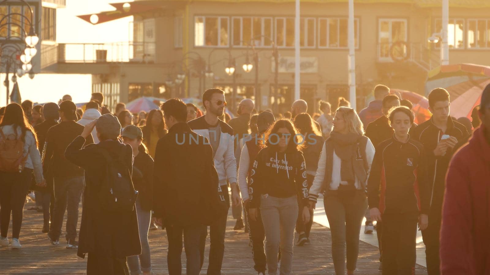 SANTA MONICA, LOS ANGELES CA USA - 19 DEC 2019: Many multiracial people walking on pier. Pedestrians walk, overcrowded seafront promenade. Crowd in golden sun light on broadwalk, sun rays over heads.