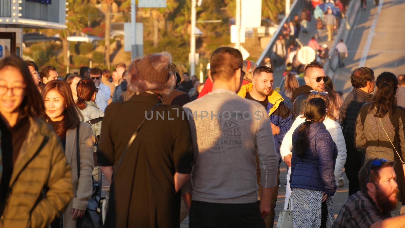 SANTA MONICA, LOS ANGELES CA USA - 19 DEC 2019: Many multiracial people walking on pier. Pedestrians walk, overcrowded seafront promenade. Crowd in golden sun light on broadwalk, sun rays over heads.