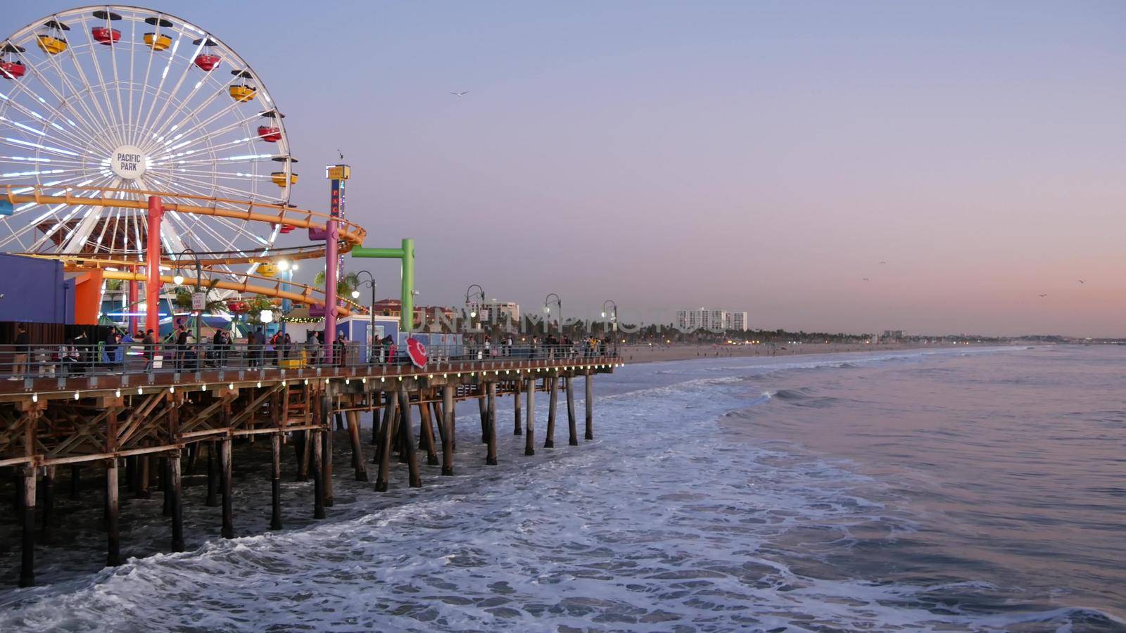 SANTA MONICA, LOS ANGELES CA USA - 19 DEC 2019: Classic illuminated ferris wheel in amusement park on pier. California summertime beach aesthetic, ocean waves in pink sunset. Summertime iconic symbol by DogoraSun