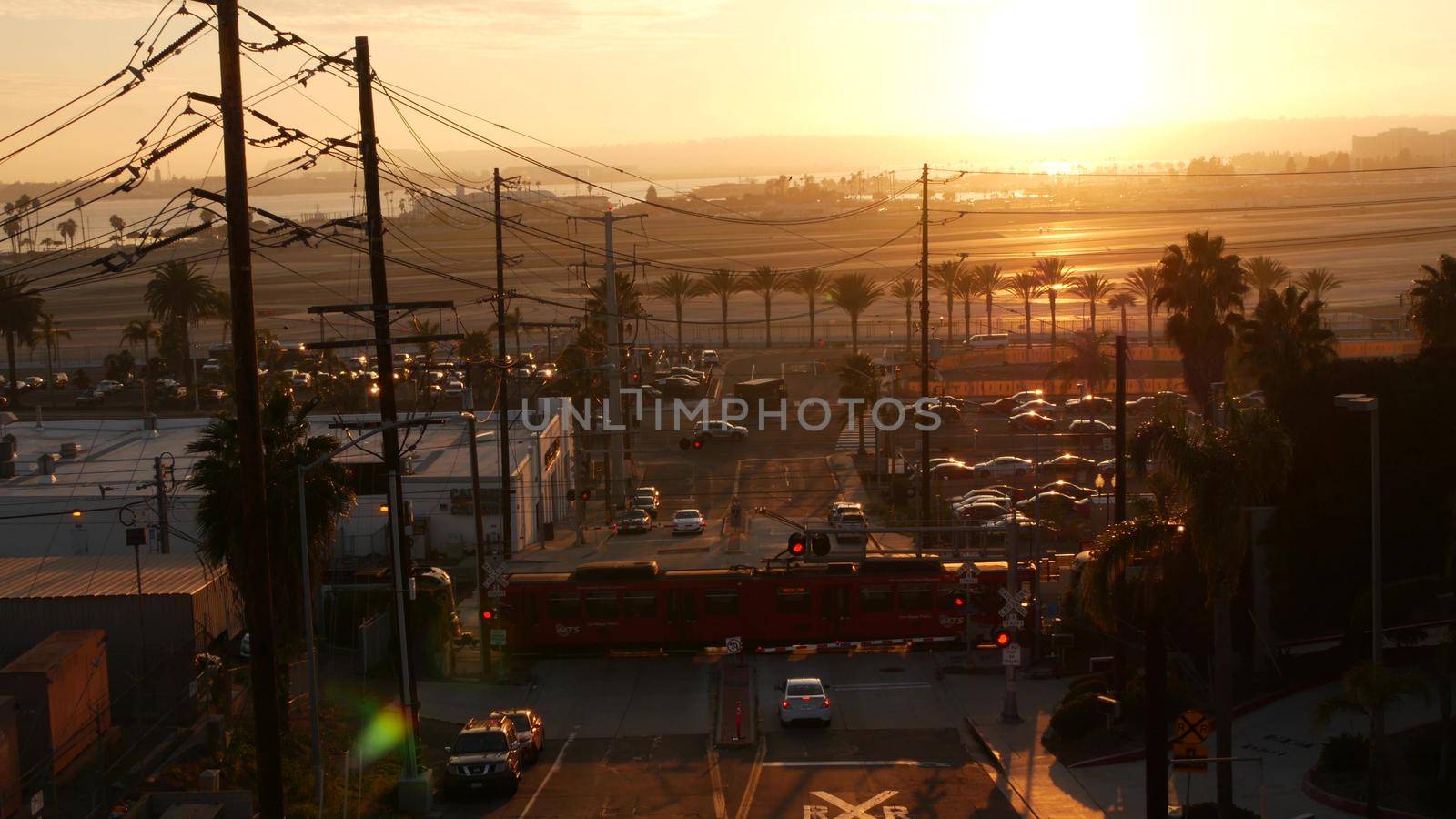 SAN DIEGO, CALIFORNIA USA - 15 JAN 2020: Level crossing near Lindbergh field international airport. Railroad red tram and runway at sunset. MTS Trolley rail transportation, cars and railway transport.