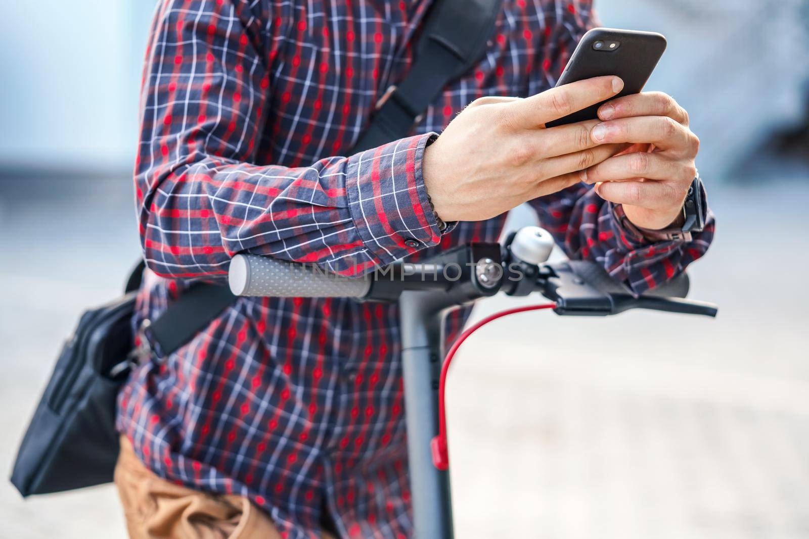 Young man wearing shirt leaning on electric scooter handlebar, holding mobile smartphone in his hands, closeup detail.