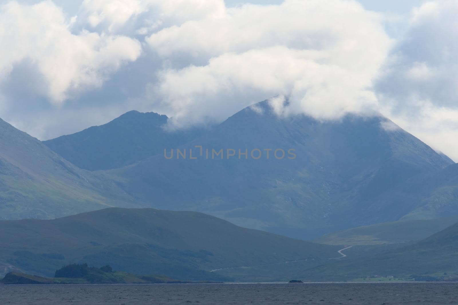 Distant road over the water winds towards a huge rocky mountain on a scottish island. Bright sunlit clouds cover the top of the mountain. Landscape.