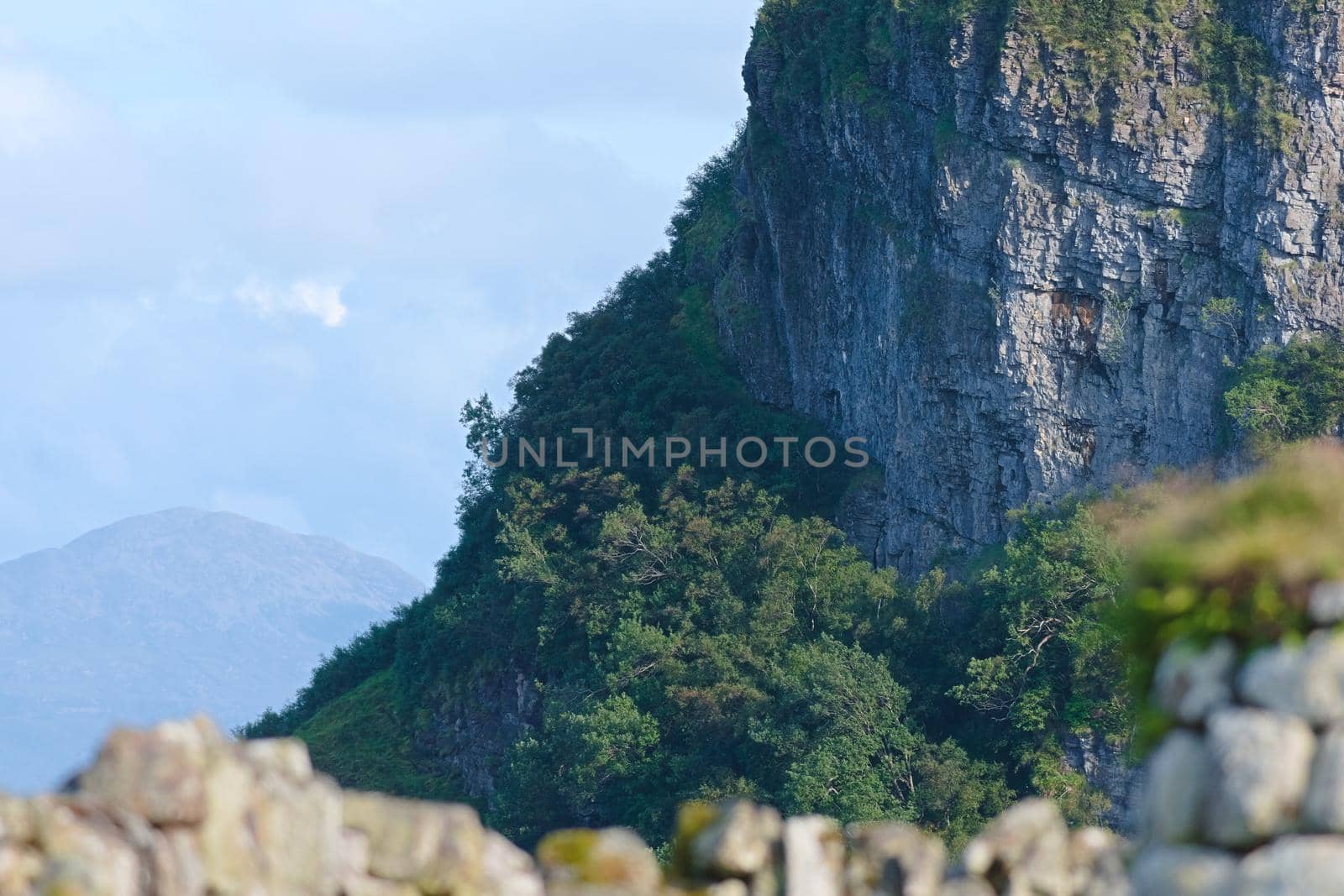 Steep jagged cliffs and distant mountains lit by evening sunlight and covered in wild trees and undergrowth. Out of focus dry stone wall foreground.