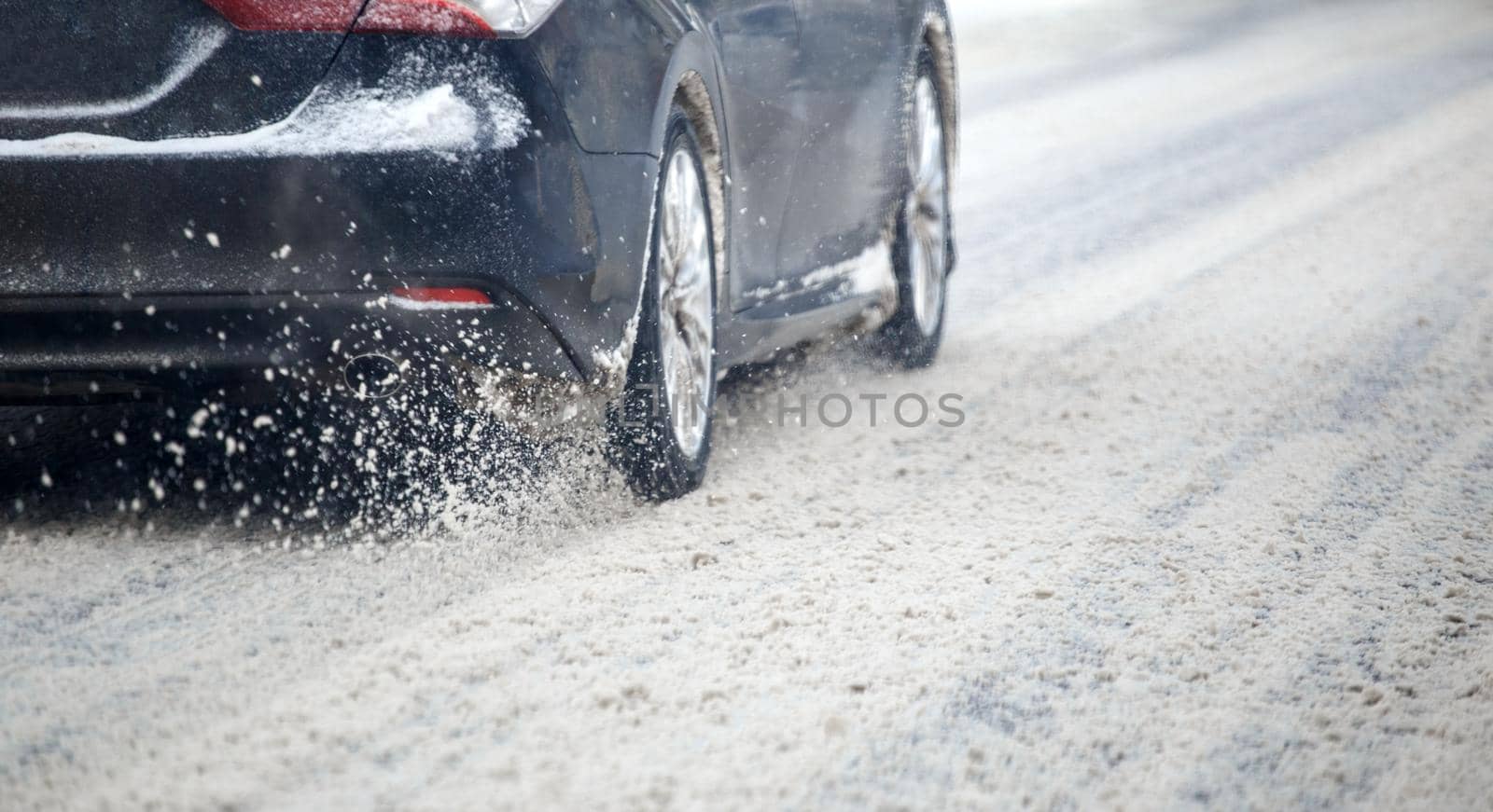 Road snow flinders flow from wheels of black car moving fast in daylight city with selective focus. Car moving on asphalt road after heavy winter snow storm.