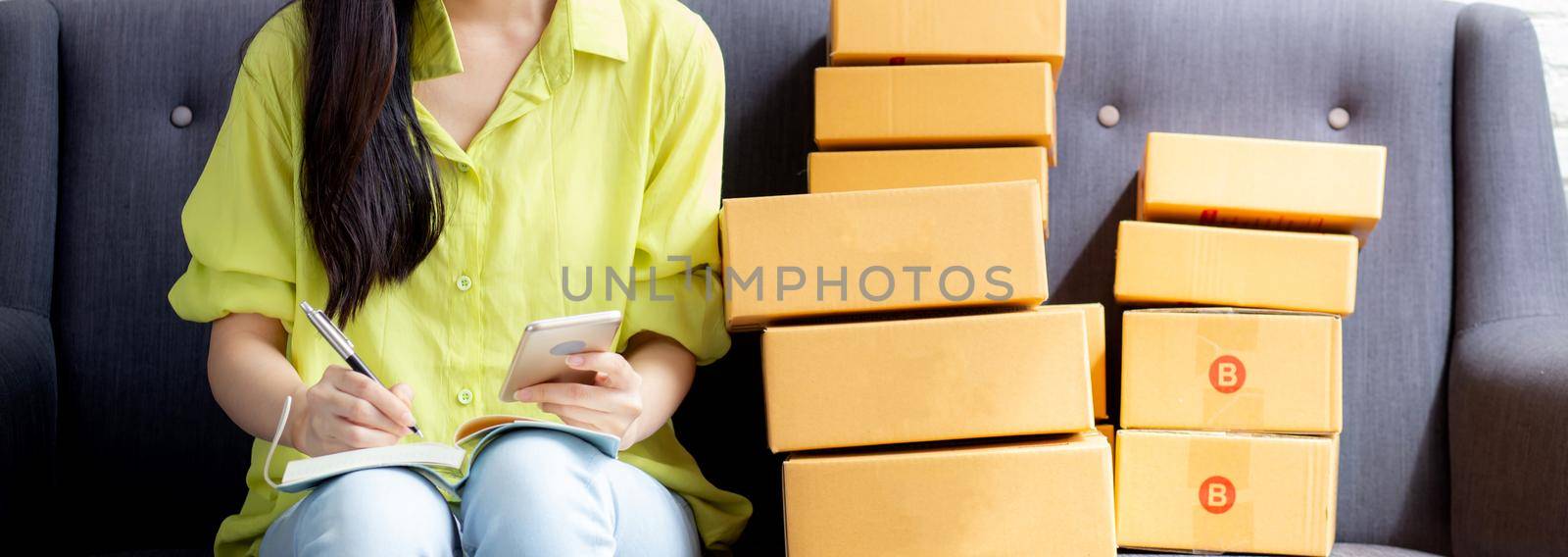 Young asian woman is entrepreneur looking phone and writing order of customer with online on notebook, online shopping, logistic and delivery, female is seller working on sofa, business SME.